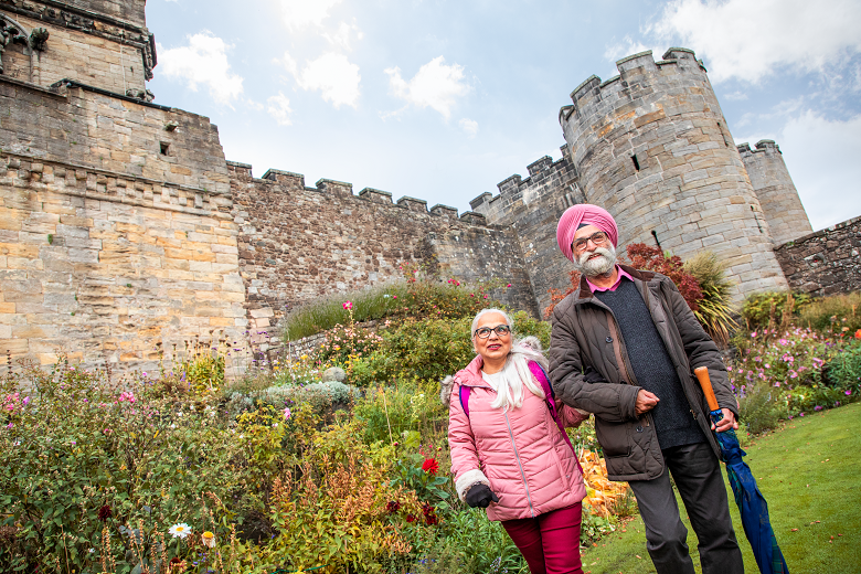 Two visitors wlaking through a castle garden, with walls and ramparts towering above them