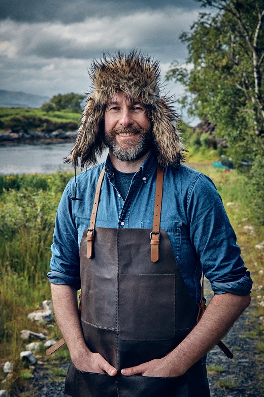 The Hebridean Baker poses outdoors wearing a wooly hat and leather apron 