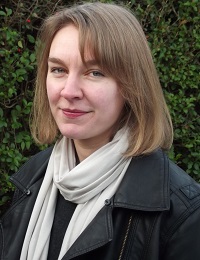 A woman with bobbed hair looks at the camera. She wears a leather jacket and light-coloured scarf.