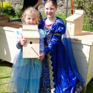 Two little girls in princess dresses hold a wooden bird box. They're outside in a garden and there is a trestle table behind them.