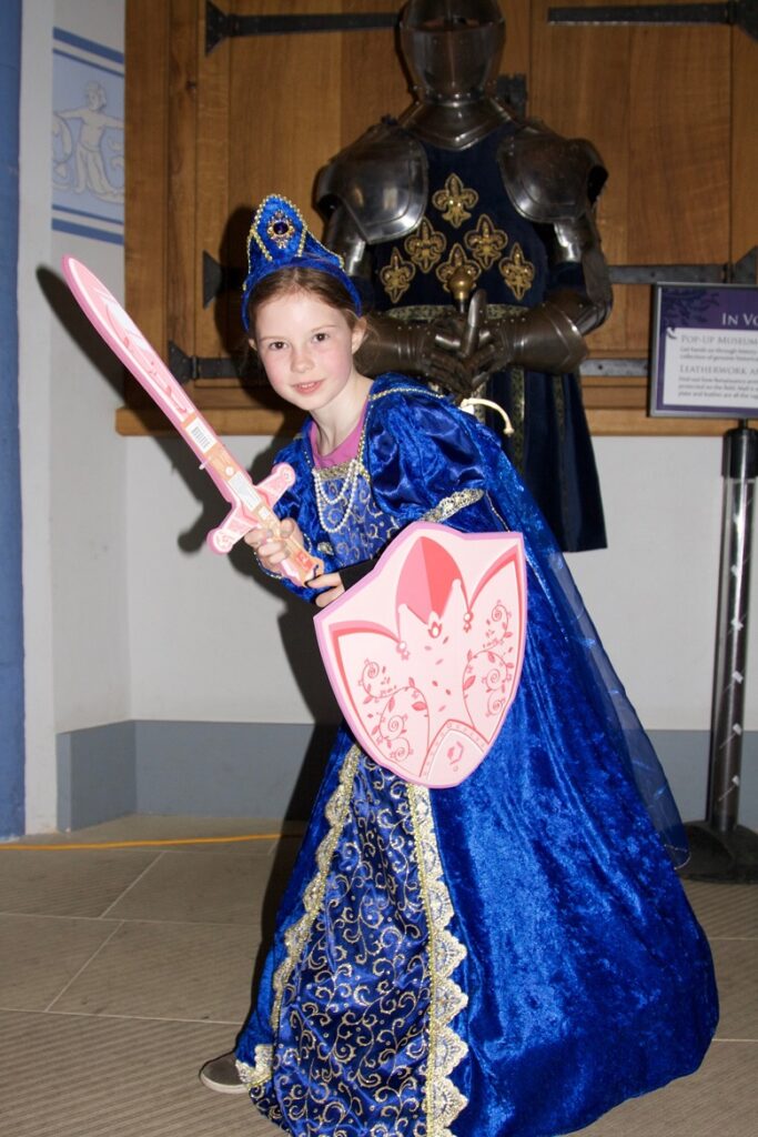A young girl wearing a blue Renaissance dress poses with a toy sword and shield in front of a suit of armour