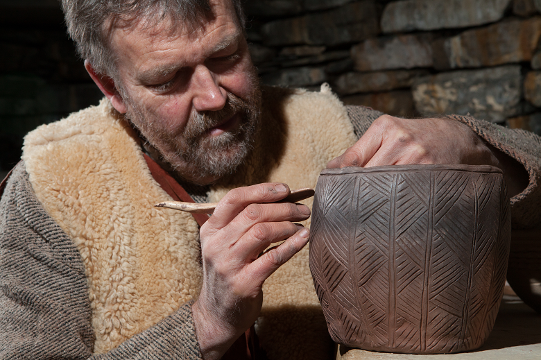 A bearded man wearing a sheepskin waistcoat over a woven jumper uses a tool to scratch a geometric design into a clay pot.