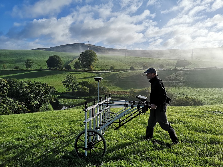 A man in a baseball cap pushed a large frame on wheels over some grassland. Behind him is a beautiful rural landscape of rolling hills on a sunny day.
