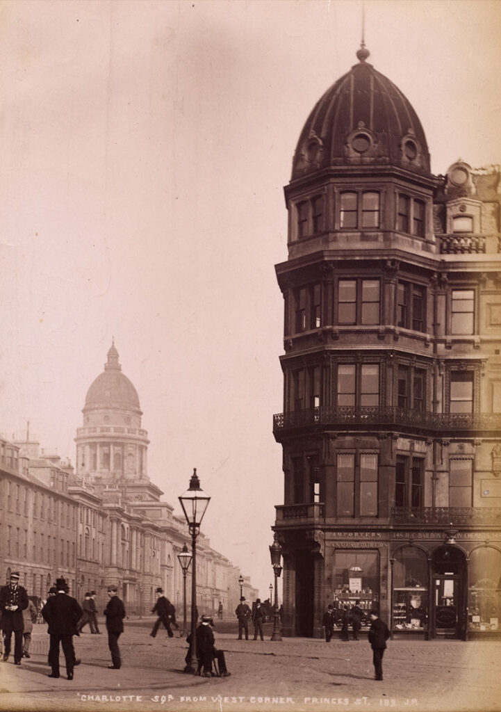 archive photo of the west end of princes street showing a building in a Victorian style with men in Victorian dress in the street.