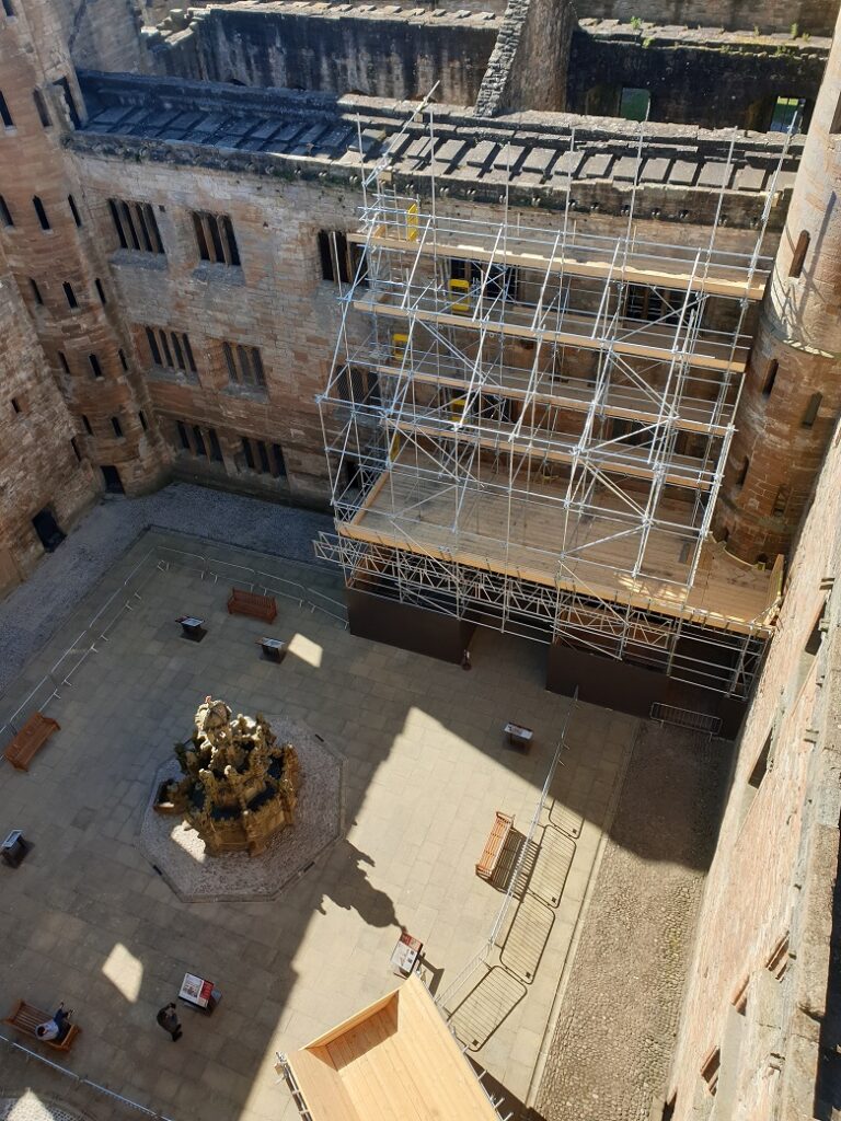 view of the courtyard at Linlithgow Palace taken from a high vantage point. There is some scaffoding in one corner and low barriers encircle the courtyard. Visitors sit on benches below.