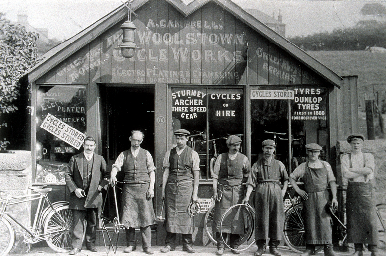An archive photo of the staff of a small cycle works posing in front of the premises. The men wear flat caps and overalls or aprons, and are holding various bicycle parts.