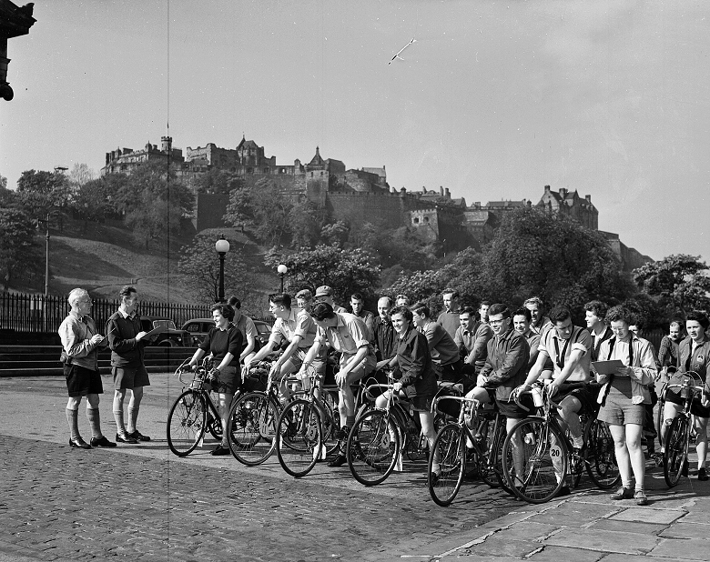 A large group of cyclists line up on a cobbled Edinburgh street, preparing to start a long tour or race. Three officials with clipboards and stop watches stand beside them. The imposing bulk of Edinburgh Castle dominates the background. 