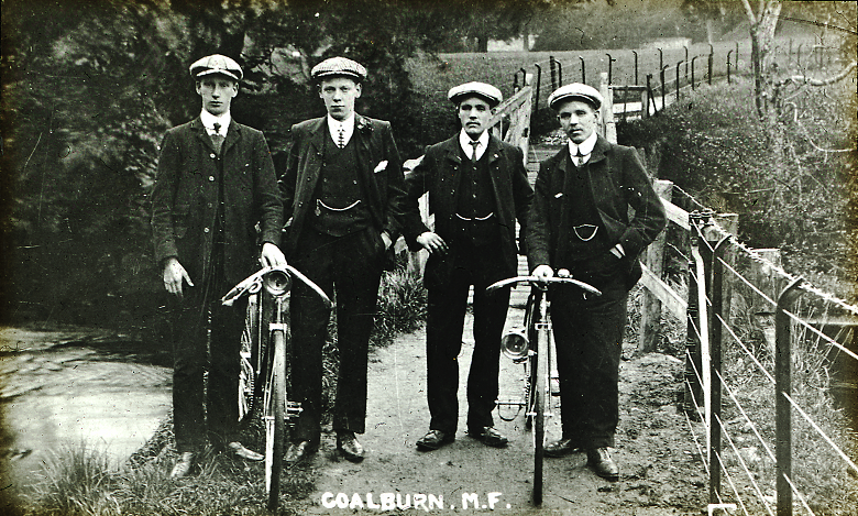 An archive photo of four men in caps posing with two safety bicycles in front of a narrow bridge