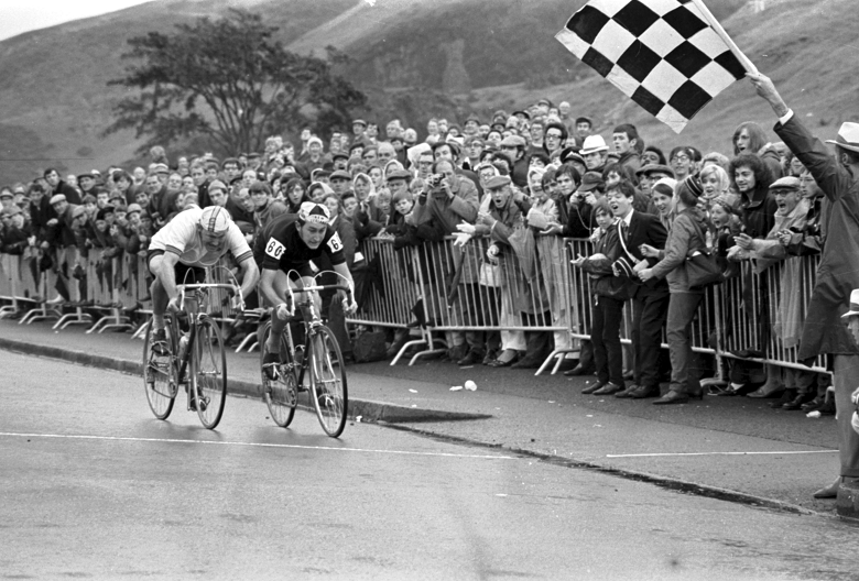 A black and white archive photo of two male cyclists racing towards the chequered flag at rhe finish line of a road cycling race, while crowds behind a barrier look on.