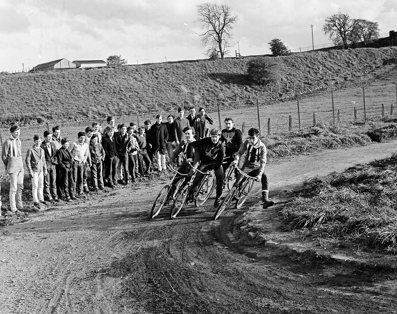 A crowd of young people look on as cycle speedway racers skid around a corner on a dirt track. The riders are teenage boys who wear distinctive team jerseys, one is striped and the other has a large star on the front,