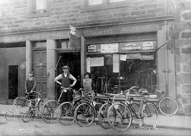 An archive photo of a man, a woman and a boy posing in front of a small bicycle shop on a terraced street. Bicycles of various makes and sizes are lined up on the pavement. 
