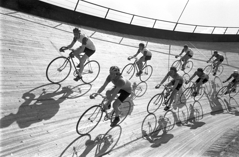 Black and white archive photo of athletes cycling on a velodrome track. The riders and their bikes cast atmospheric shadows onto the track.