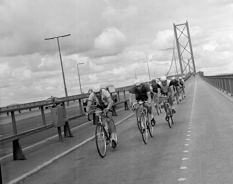 A black and white archive photo of a group of riders crossing the Forth Road Bridge during a cycling race. The suspension bridge's distinctive towers can be seen behind them.