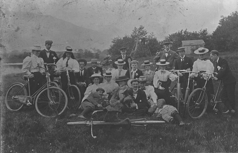 A slightly damaged archive photo of a well-dressed cycling group on a countryside trip. The group is made up of men and women and some are holding instruments including a guitar or banjo. 
