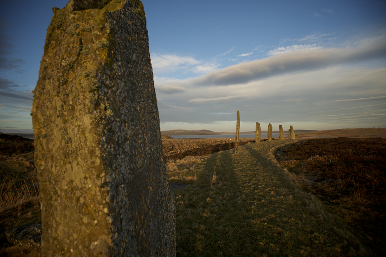 A close-up photo of an ancient, moss-covered standing stone at the Ring of Brodgar. In the background there are more large standing stones, which are casting shadows across the hetaher and grass,