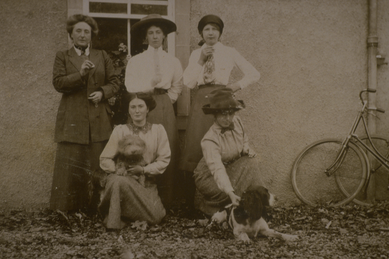 A sepia photograph of a group of female cyclists posing wth two dogs. Their bicycles can be seen leant against a wall beside them. 
