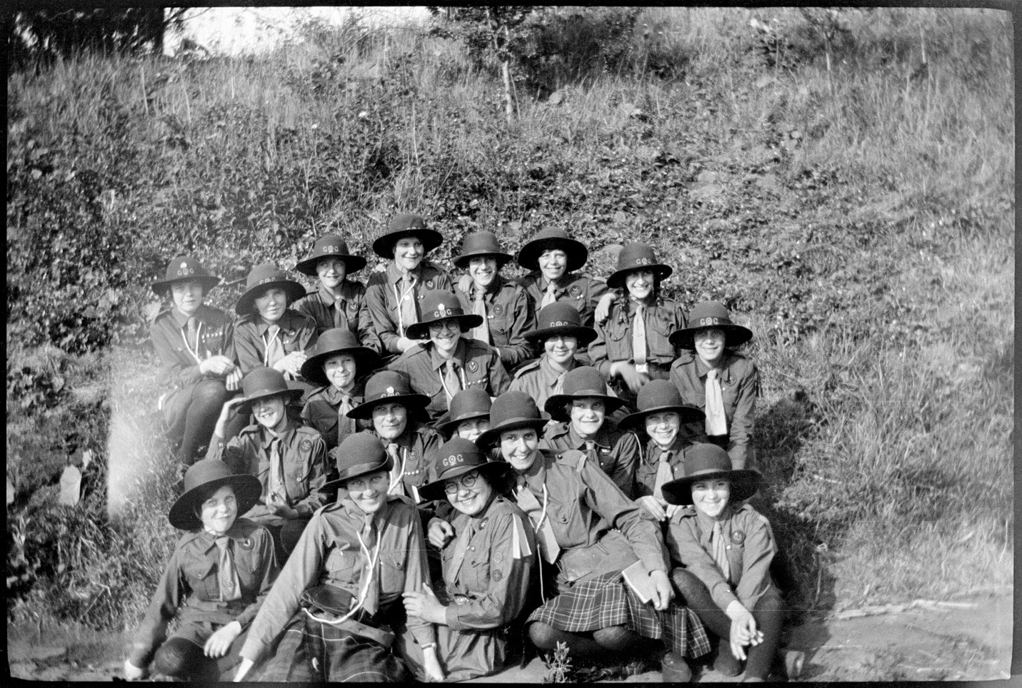 A black and white archive photo of a Girlguiding group wearing helmets and uniforms posing for a photograph sitting in greenery. 