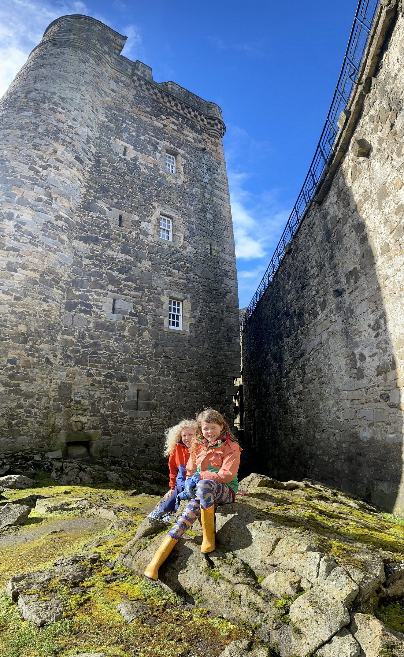 Two children sitting on rocks in front of Blackness Castle wearing wellies. 
