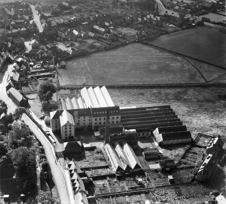 A balck and white aerial photo of a large four-story mill building surrounded by ancillary buildings. the streets and houses of a small town can be seen in the background. 