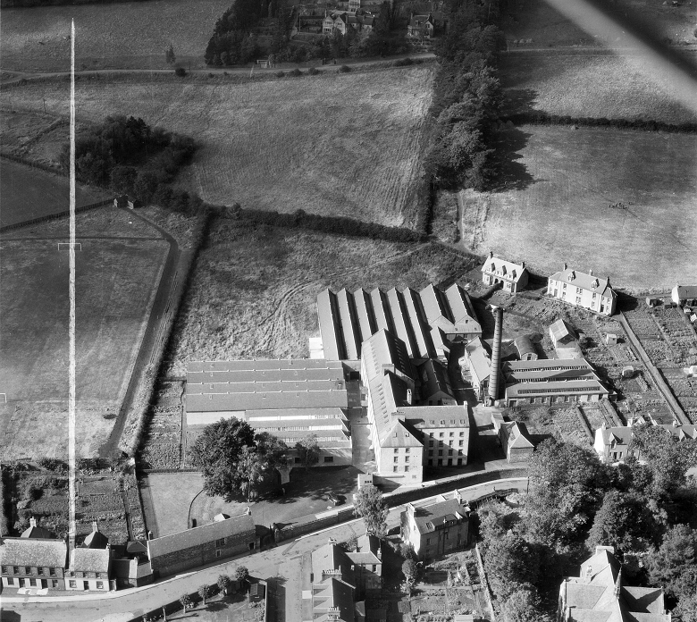 A black and white archive photo taken from an aeroplane showing a large mill complex on the edge of a small town. Beyond the mill there is rolling farmland and woods. 