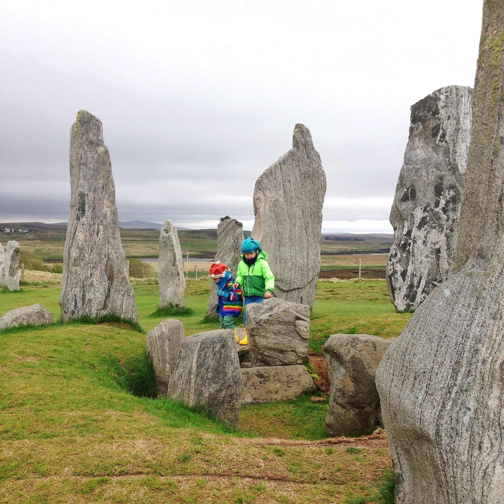 Two children walking between the Calanais Standing Stones. The children are wrapped up warm in colourful rain jackets and wellies and hats. It looks like it's about to rain.