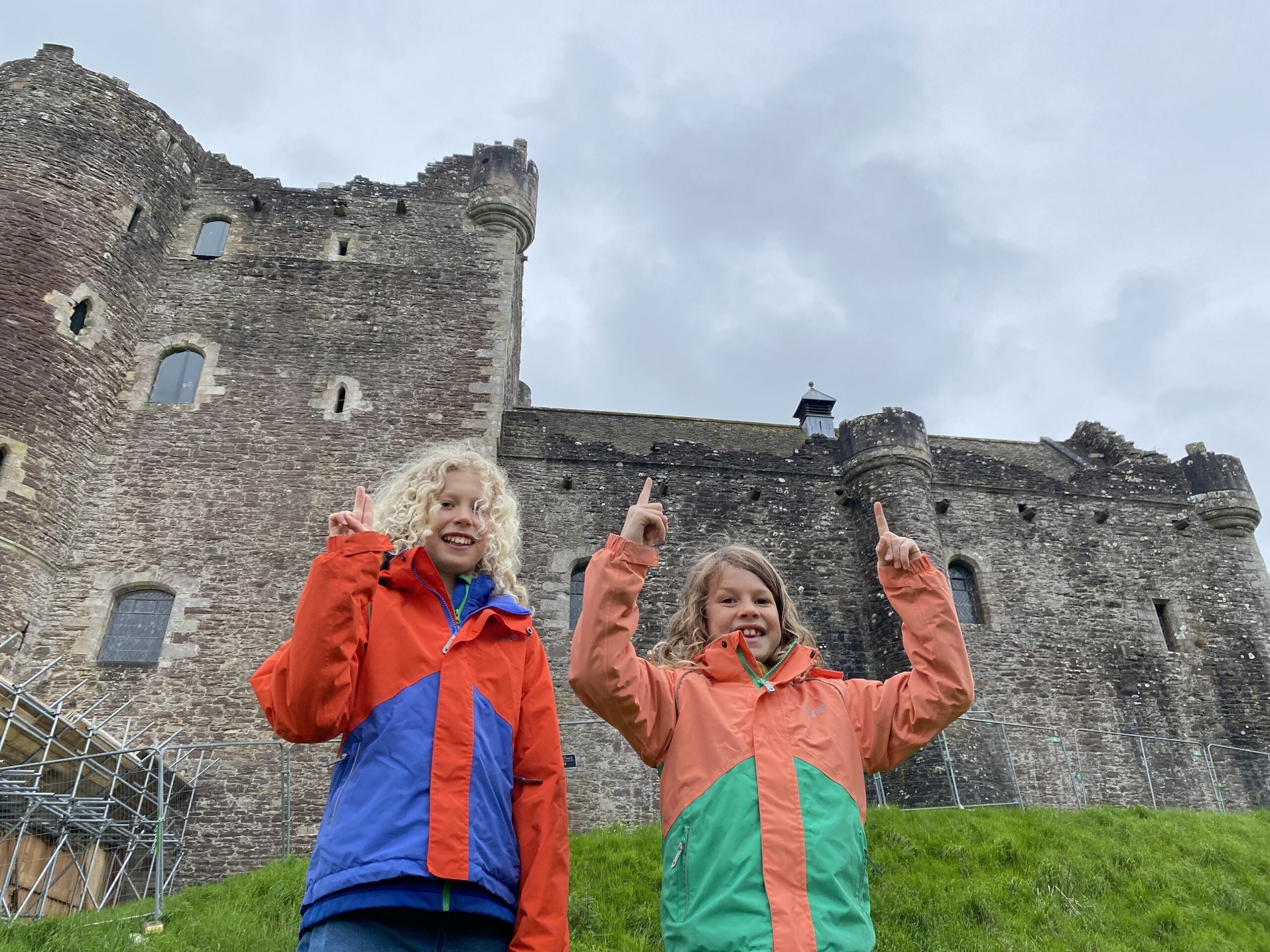 Two children standing in front of Doune Castle. They are smiling at the camera and are pointing at Doune Castle. They have long blond hair and are wearing bright jackets with similar patterns. 
