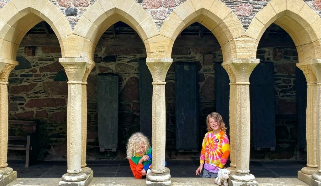 2 children with blonde long hair standing between the archways of Iona Abbey. They are wearing bright, colourful jumpers.