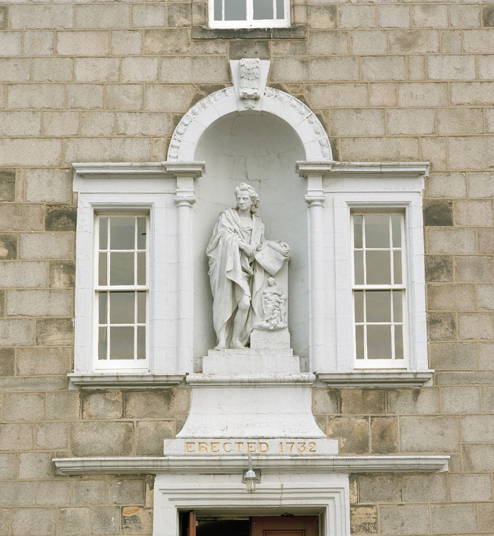  A white marble statue of a man depicted wearing robes and holding a parchment or scroll. The statue is set into the front of a stone building and flanked by two windows. An engraving beneath it tells us it was erected in 1732.