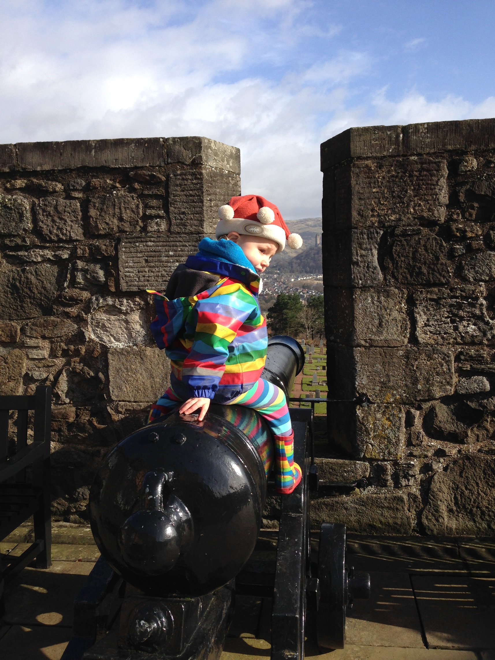A young child wearing a Santa hat and a rainbow rain jacket with matching leggings sitting on a canon at Stirling Castle. 