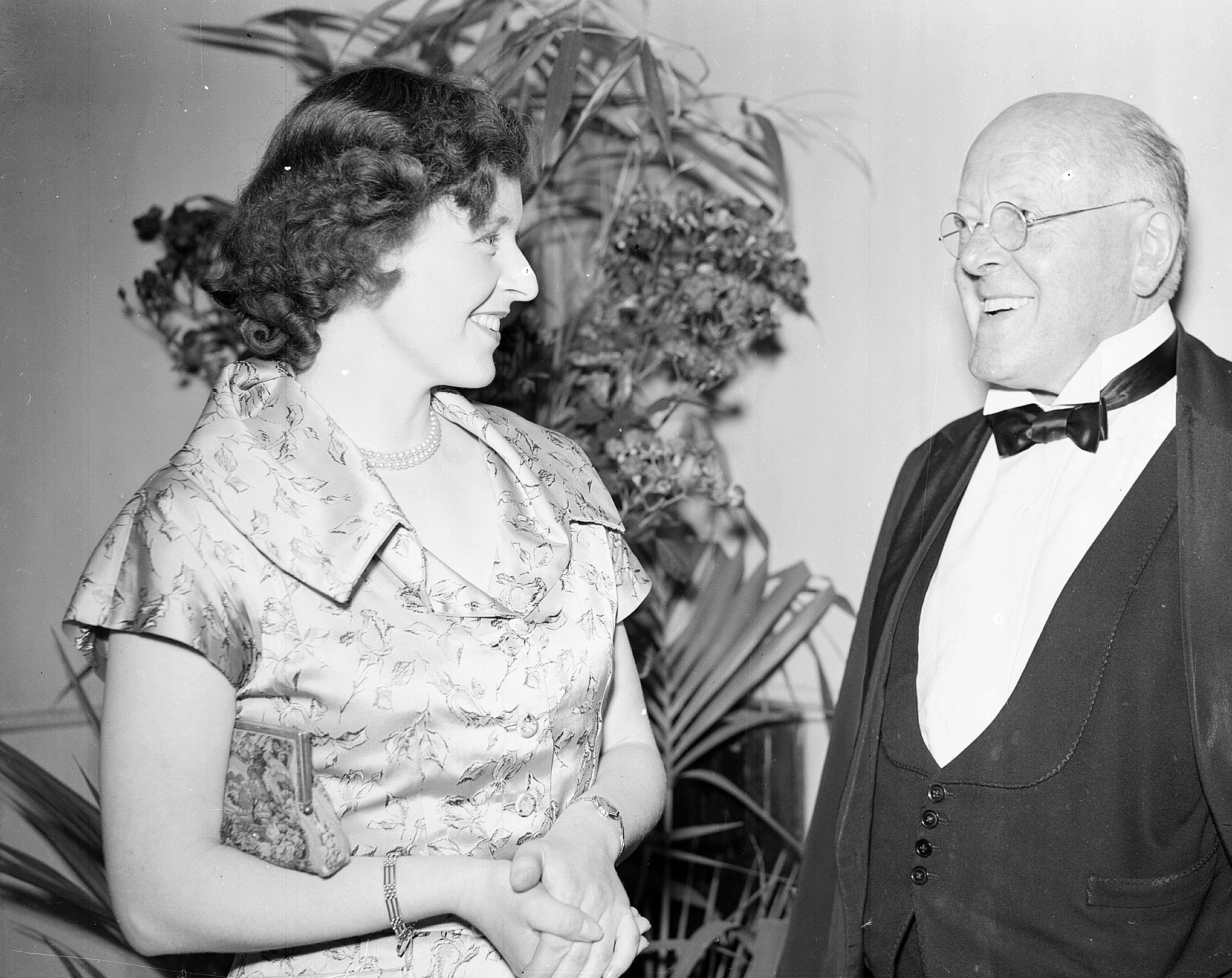 A black and white archive photo of a young woman wearing a patterned, collared silk dress and a bracelet and an older man with glasses wearing a tuxedo. They are smiling at each other. 
