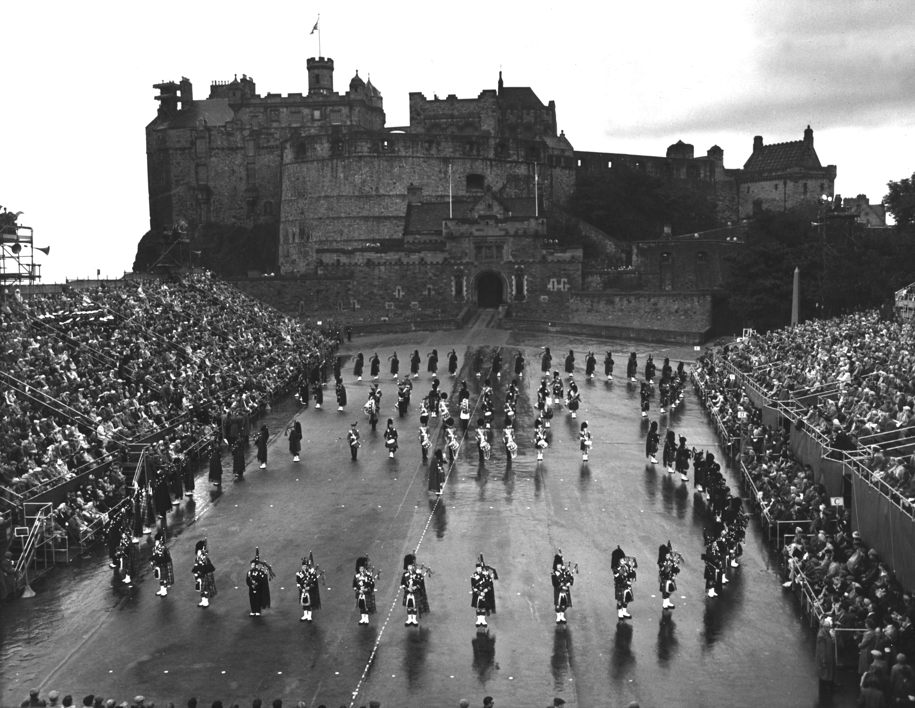  black and white archive photo showing a performance of the Edinburgh Military Tattoo. You can see the stands are very busy with spectators looking at a scene of the military parade against the backdrop of Edinburgh Castle.
