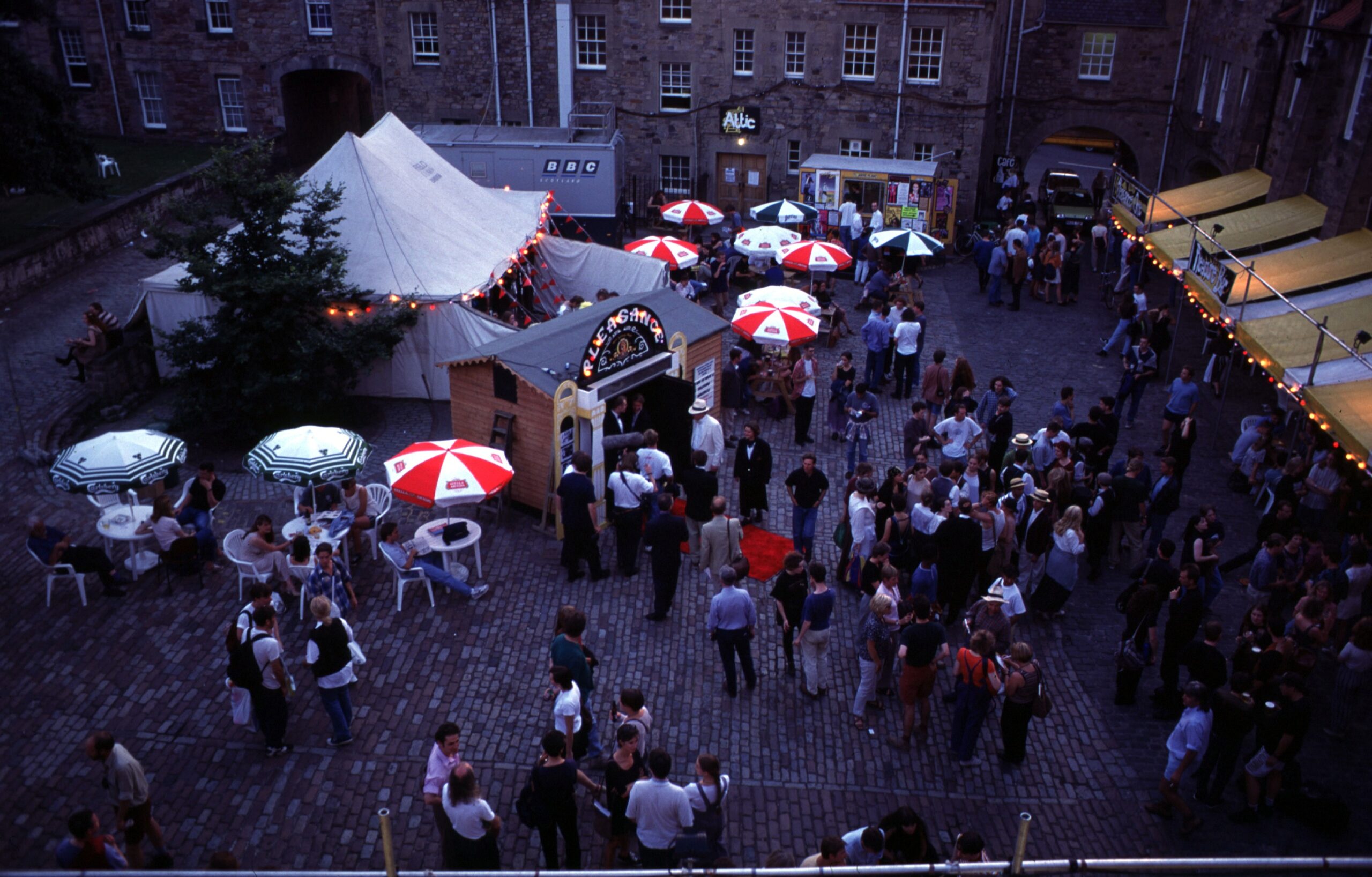 A view overlooking the Pleasance courtyard during the Edinburgh Festival where the smallest theatre was located. There are crowds gathering in the courtyard and the small stands.