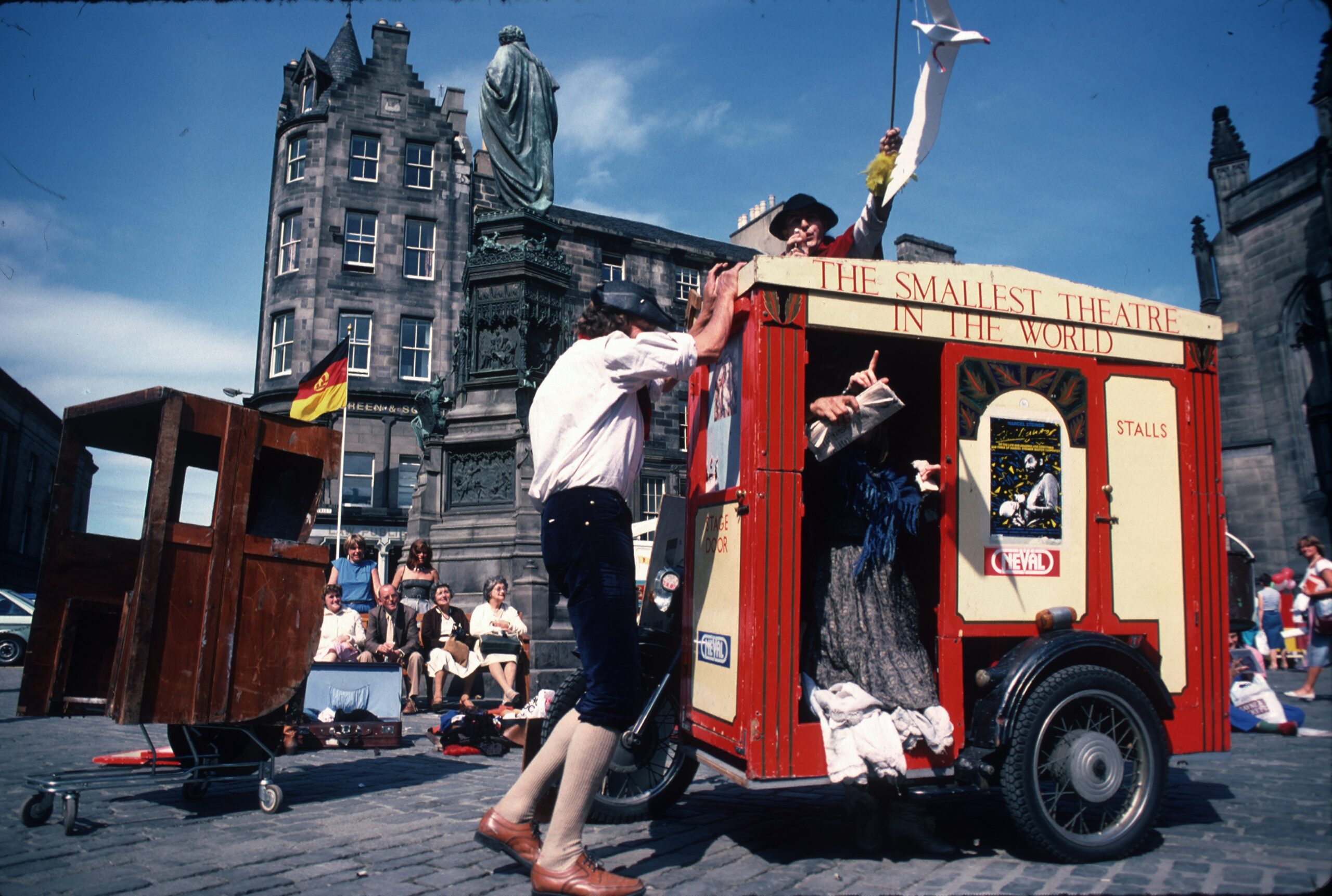 A colour archive photo of the bike with the smallest theatre in the world on a street in Edinburgh. There is a man standing behind the theatre looking over it. Another man is standing next to it. 