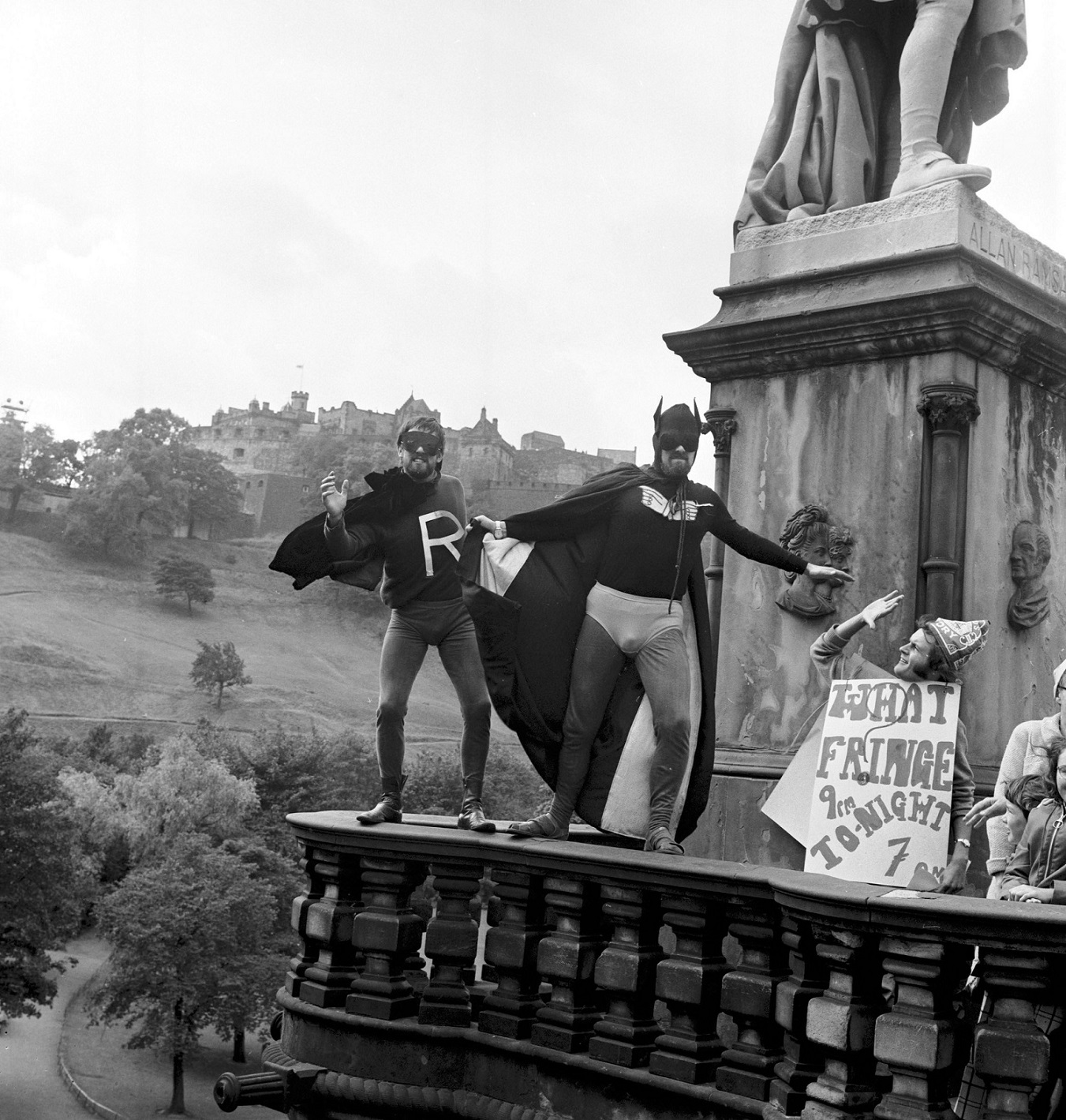 A black and white archive photo from the Edinburgh Festival. You can see two people standing on a statue in Princes Street Gardens dressed as Batman and Robin. Batman is pointing to a a man holding a sign saying "What Fringe"