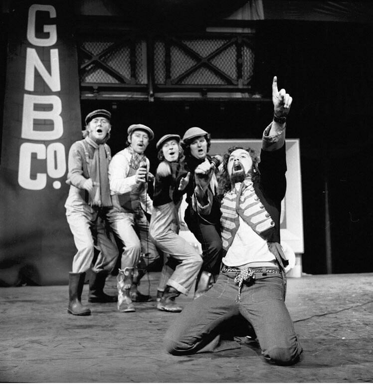 A black and white archive photo from the stage performance of the The Great Northern Welly Boot Show. Billy Connolly is singing into a microphone, kneeling on the floor and pointing up. Four male actors in costumes are standing behind him singing as well. 