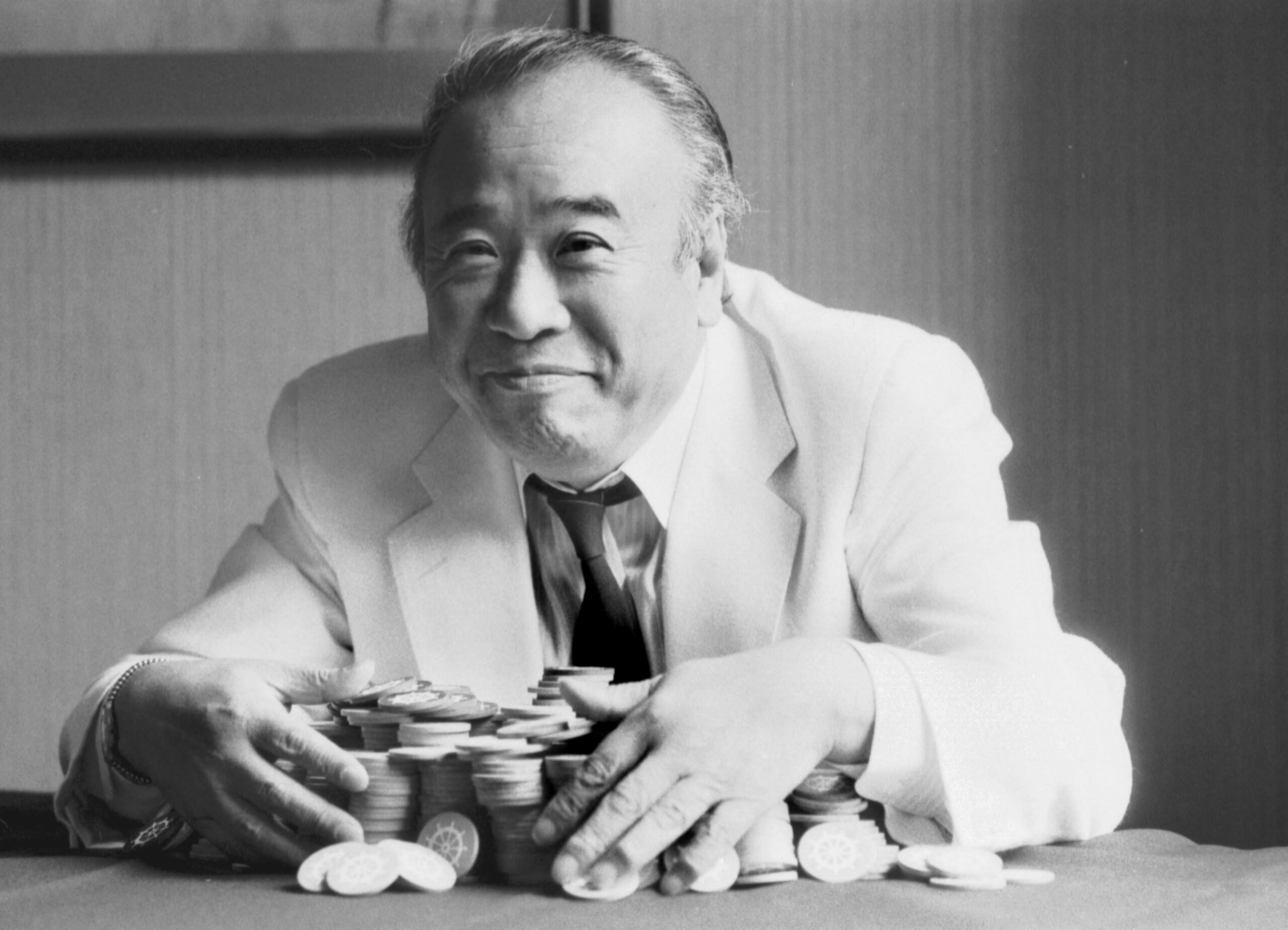 A black and white photo of an older Zenya Hamada. He's wearing a white suit and has a pile of gambling chips in his arms. He's smiling at the camera. 