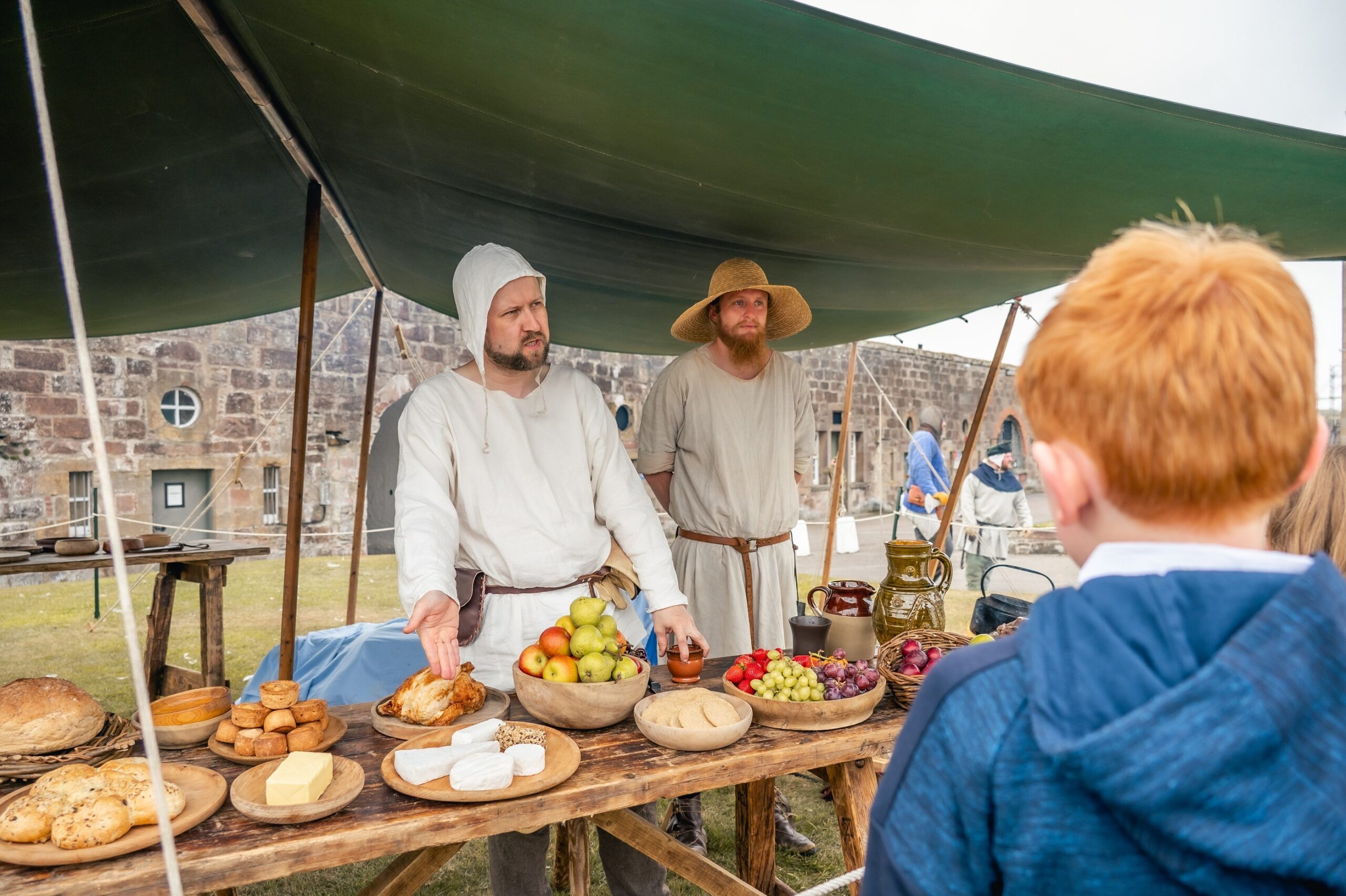 Two men dressed as medieval farmers standing at a market stand selling bread, fruit and cheese at the Celebration of the Centuries event at Fort George. 
