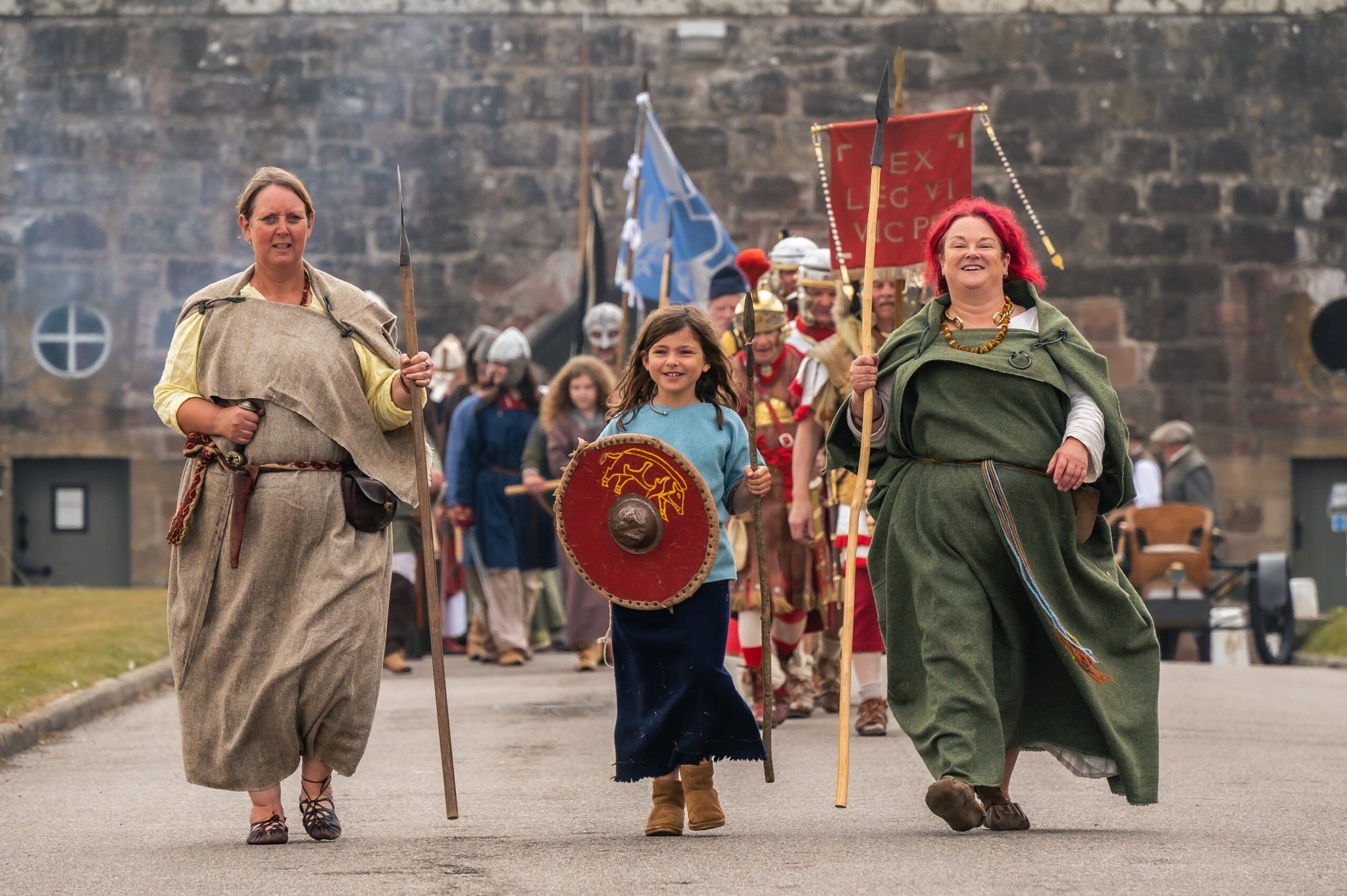 Two women and a young girl dressed in medieval clothing smiling towards the camera. The young girl is holding a shield and a spear. The women are holding spears as well. 