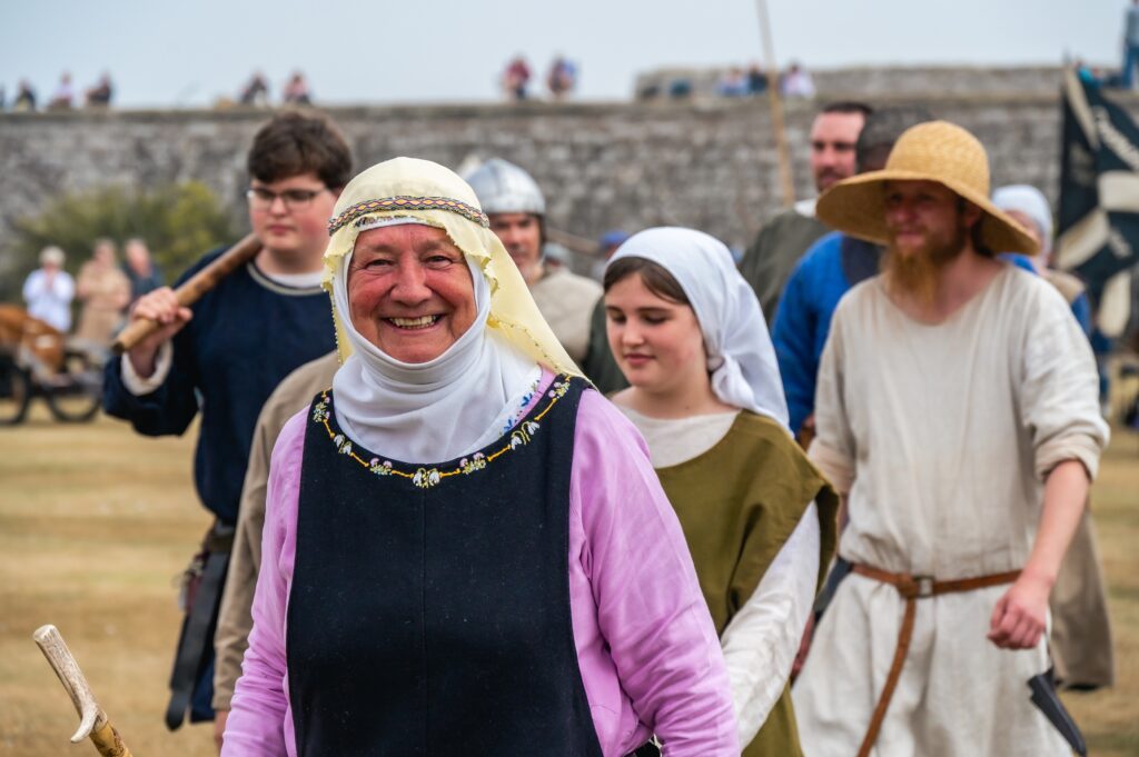 Six re-enactors at the Celebration of the Centuries 2022 event at Fort George. The woman that is most promintent in the photo is wearing a medieval headscarf, shirt and necklace and smiles at the camera. In the background you can see a young girl and a farmer dressed up as well.