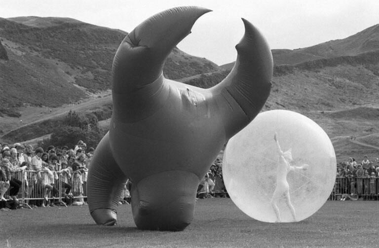 A black and white archive photo from the Fringe Sunday at Holyrood Park as part of the Edinburgh Festivals. You can see a person in a zorbing ball and a person in a blow-up suit in an odd shape.