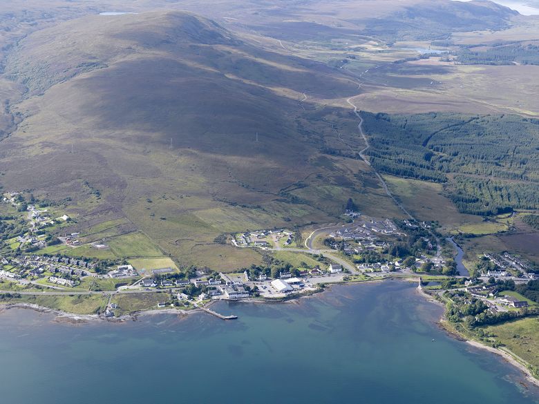 An aerial view of an island village centred around a bay. A large expanse of moorland and forest is on the hillside begind the village.