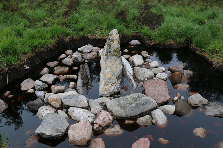The remains of a prehistoric burial cairn, partially submerged in water. A manmade structure resembling a small standing stone can be made out. 