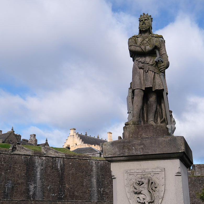 King Robert I depicted in a full length statue on top of a plinth. He wears medieval dress, including a tunic, chain mail and armoured gloves. He wears a crown and has a beard and carries a sword.