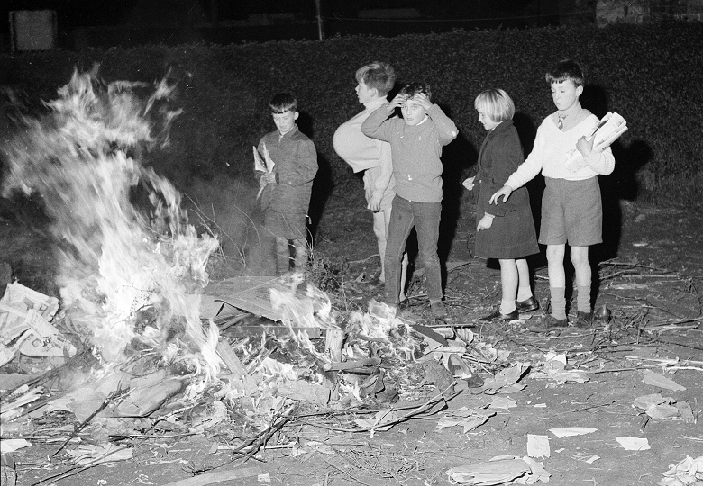 Five children stand around a large bonfire. It's the 1960s and the children are dressed in the fashions of the day. Scraps of paper blow in the foreground. 