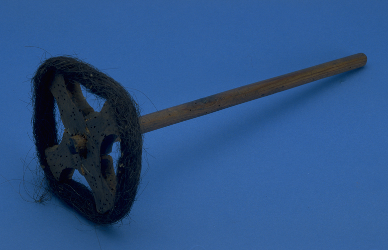 An old kitchen utensil. A long central wooden spindle ends in a cross which is wound around with horse hair.