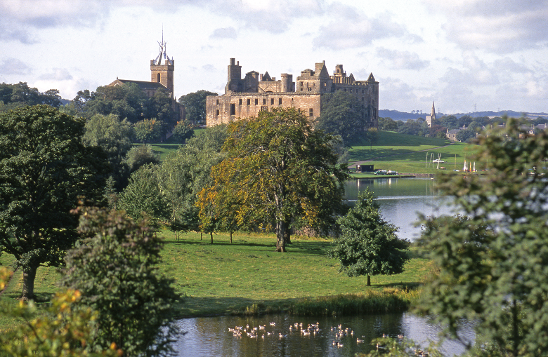 Distant view of the ruins of Linlithgow Palace. In the foreground is a picturesque loch with a flock of geese on the water. It is surrounded by parkland with mature trees. THe ruins of the palace sit on the hill above, the steeple of a church is behind.