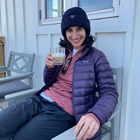 A woman wearing warm and sporty outdoor clothing sits outside a cafe drinking a coffee.