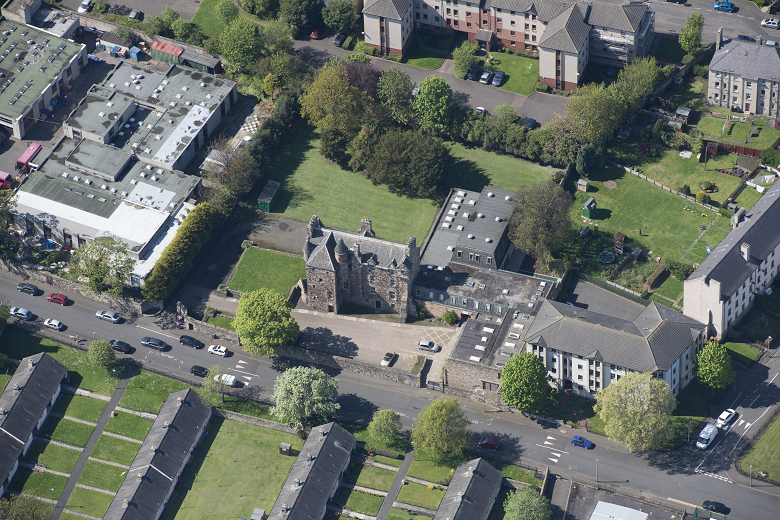 Aerial view of the house showing a car park to the front. Some small gardens remain immediately around it. Flats and terraced housing are nearby.