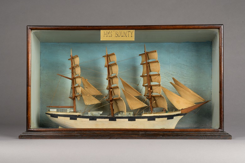 A model of the ship HMS Bounty on display in a glass case. It is a large wooden ship with three main masts and some 20 sails. 