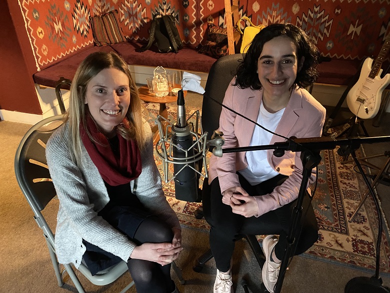 Two women smile at the camera. They are in a cosy room with decorative rugs on the walls and floor and there is a guitar behind them. A professional microphone is in front of them.
